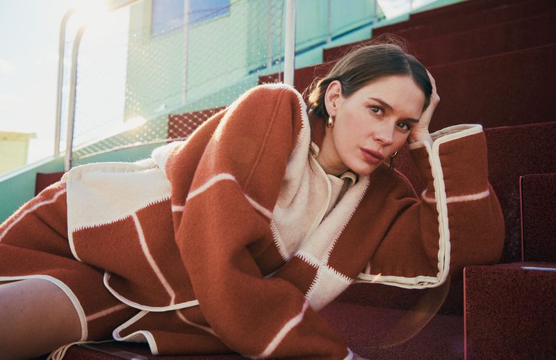 Woman in a brown and white patterned coat is lying on red stairs bathed in sunlight. The background features a window and sky.