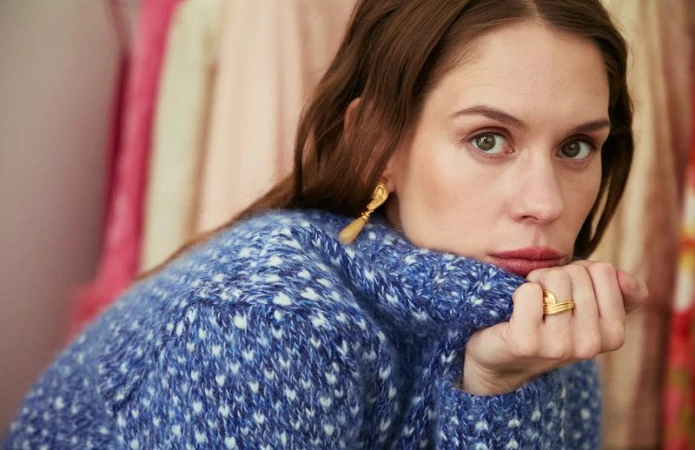 Close-up portrait of a brunette adult female wearing a blue sweater with gold earrings and rings, set against a blurred rack of clothing.