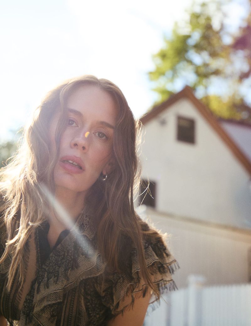Portrait of a young adult woman with wavy hair and patterned shawl outdoors, with a building and trees blurred in background.