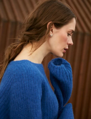 Close-up studio portrait of a young adult woman with long brown hair wearing a blue sweater, posing in front of a brown background.