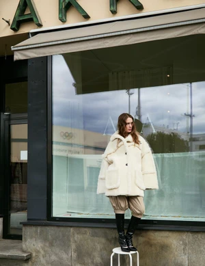 Model wearing a cream shearling jacket and tall black boots standing outside a storefront window in Vogue Scandinavia