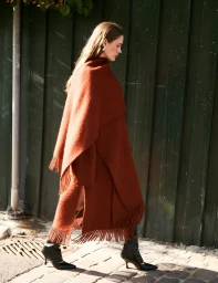 Woman walking outdoors wearing a rust-colored fringed poncho and black heeled boots against a dark wooden wall.
