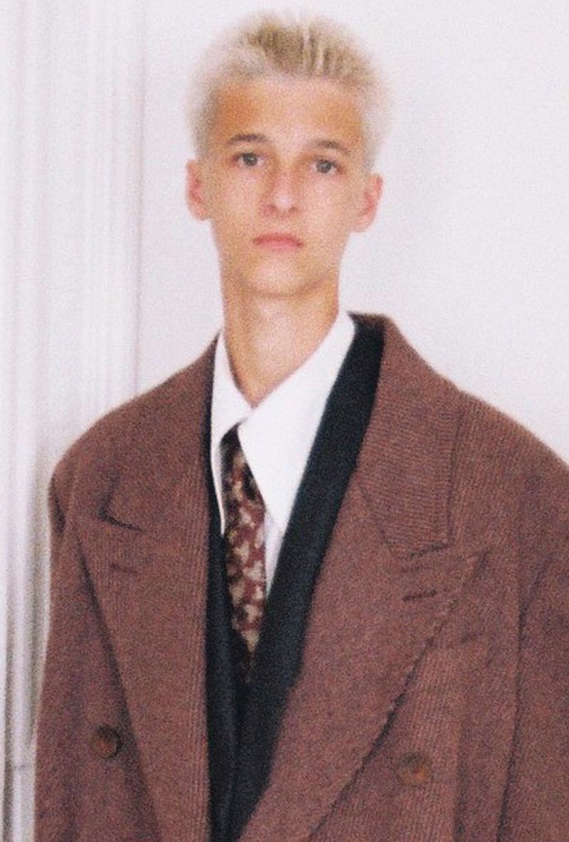 Portrait of a young adult male with blond hair wearing a brown tweed double-breasted coat, vest, patterned tie, and white collared shirt.