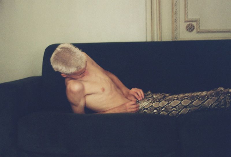 Blond-haired shirtless man with snake-print pants relaxes on a dark blue sofa in a minimalist interior setting, looking down.