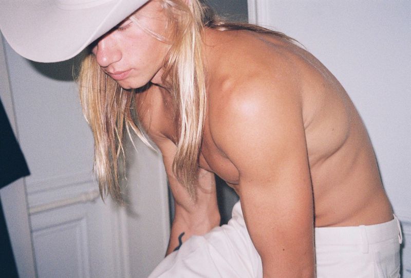 Man with long blonde hair wears a cowboy hat, looking down. Shirtless view showcases his physique against a neutral indoor background.