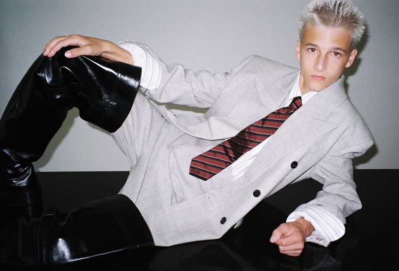 A fair-skinned young adult with spiky white hair poses in an oversized grey suit and tie with shiny black thigh-high boots on a glossy black surface.