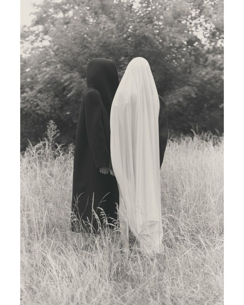 Monochrome outdoor shot of two cloaked figures in a grassy field with trees in the background, evoking a sense of mystery and artistic expression.