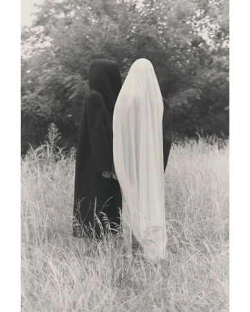 Monochrome outdoor shot of two cloaked figures in a grassy field with trees in the background, evoking a sense of mystery and artistic expression.