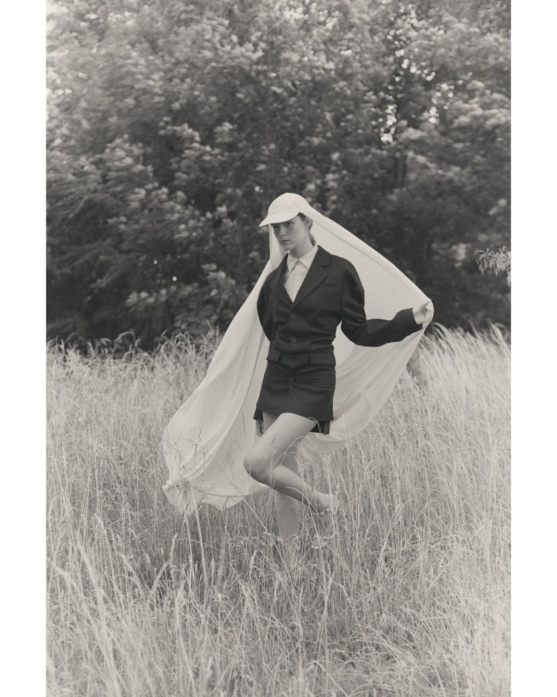 Monochrome portrait of a person in a blazer and cap standing in a field with a sheer veil, creating an ethereal, natural outdoor scene.