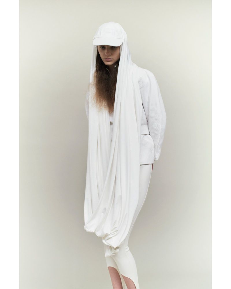 Fashion shot of a young adult woman wearing a white cap, jacket, and leggings, and a draped white shawl over her head and shoulders.