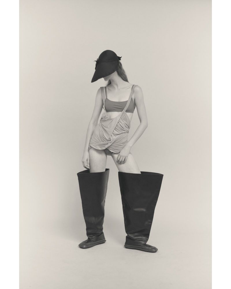 Monochromatic studio shot of a person in a hat, crop top, and oversized boots against a solid backdrop, fashion-forward, artistic.