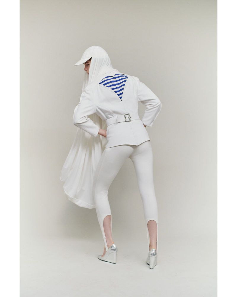 Fashion model wears white outfit with blazer, leggings, cap and wedge heels in studio setting, posing with hands on hips.