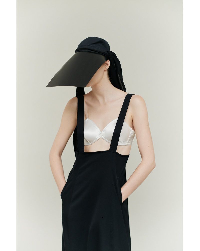 A woman poses indoors wearing a black dress, white bra, and black visor-like hat against a neutral background in a fashion studio shot.