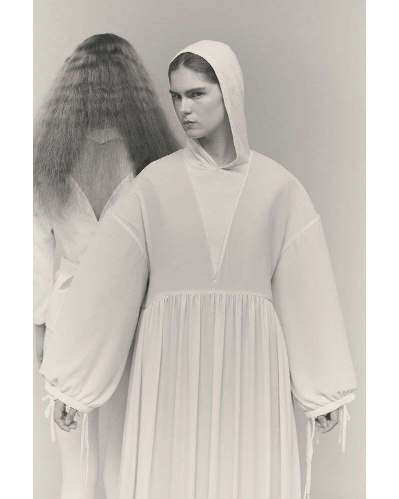 Two women in white hooded dresses pose in a studio setting. The model in the foreground faces the camera, while the other has long hair.