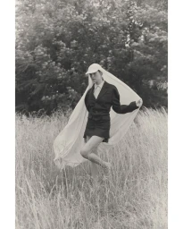 Monochrome fashion portrait of a woman in a dark tailored blazer, miniskirt, and white veiled cap standing in a tall grass field.