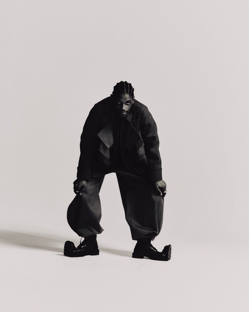 Full-length black and white studio shot of an African American male in an oversized black jacket and pants, hat in hand, and black boots.