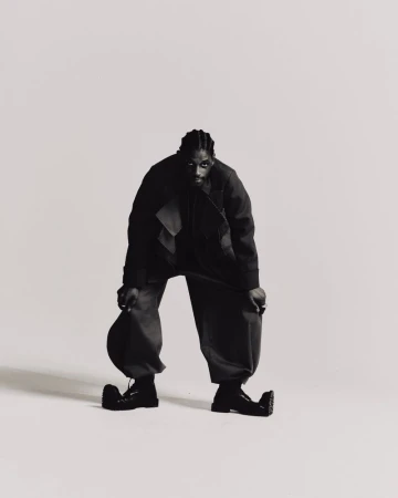 Full-length black and white studio shot of an African American male in an oversized black jacket and pants, hat in hand, and black boots.