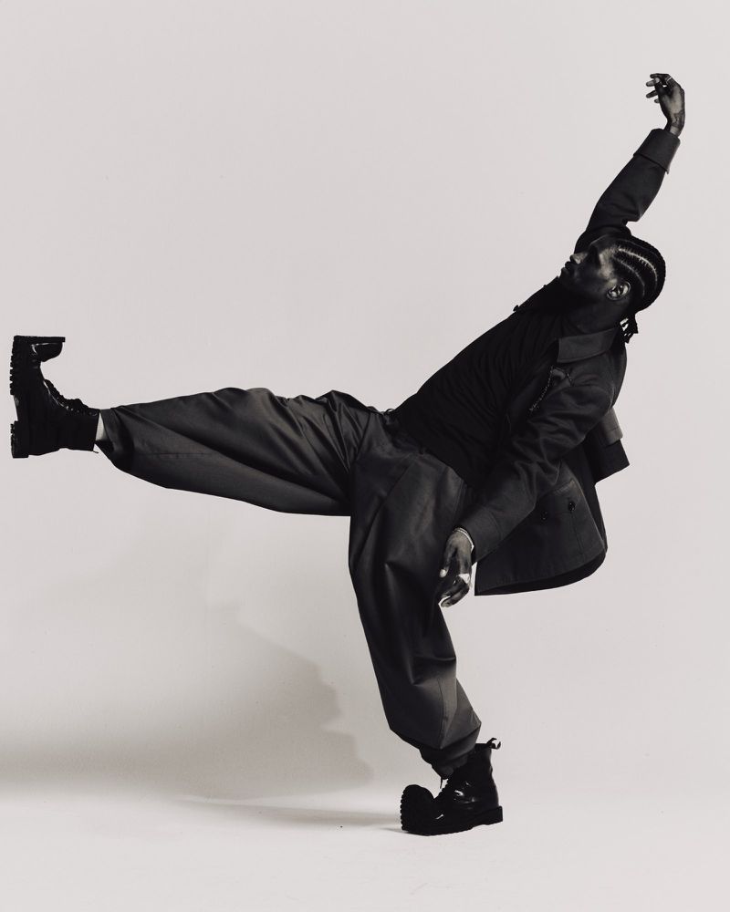 ASAP Nast in an all-black outfit with wide-leg trousers and boots poses dynamically against an off-white studio backdrop.