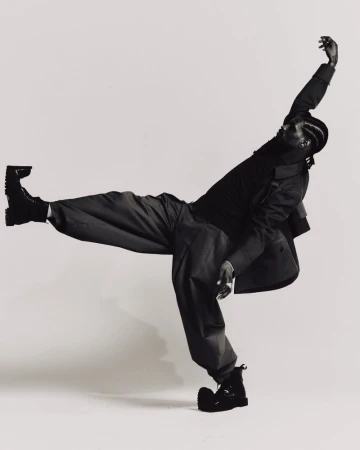 ASAP Nast in an all-black outfit with wide-leg trousers and boots poses dynamically against an off-white studio backdrop.