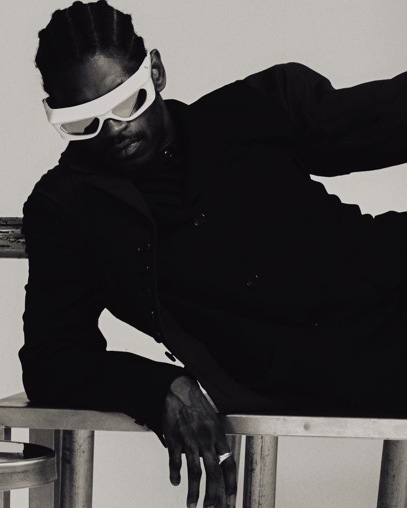 Stylish studio portrait of a Black man wearing futuristic sunglasses, a dark blazer, and striking pose on a metal chair.