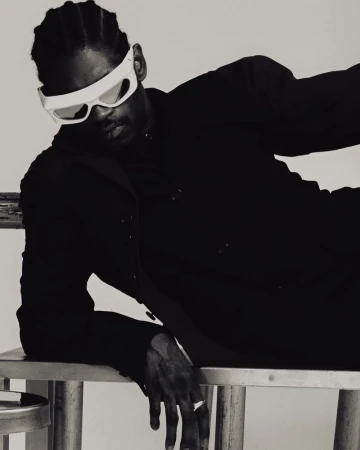 Stylish studio portrait of a Black man wearing futuristic sunglasses, a dark blazer, and striking pose on a metal chair.