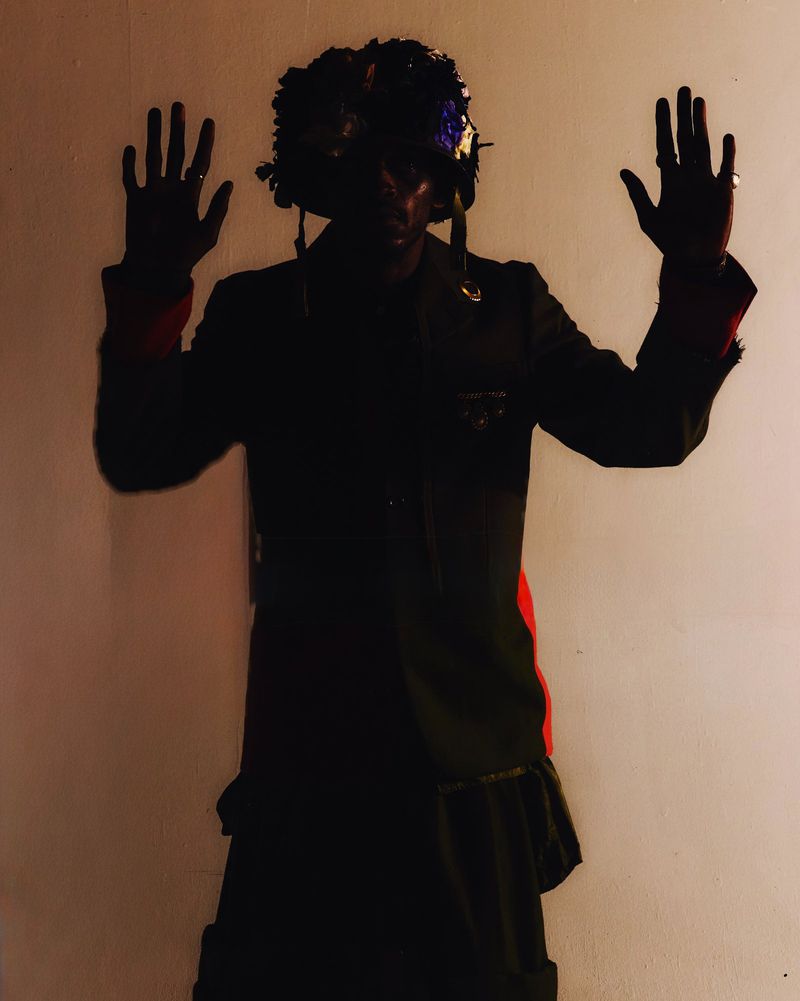 Person stands with hands up wearing a black floral hat and jacket in a dimly lit indoor studio setting, showcasing dark green and red tones.