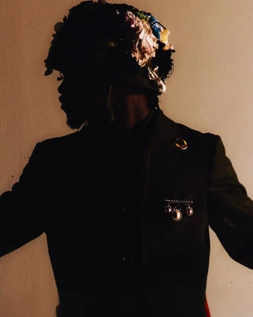 Portrait of a Black male in vintage military jacket with floral headpiece against a neutral backdrop, creating a dramatic and artistic silhouette.