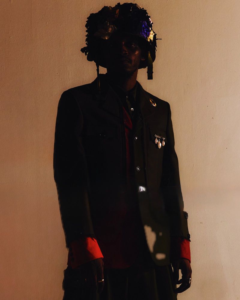Individual in dark jacket with floral headpiece poses in dimly lit setting, creating a visually striking and unique fashion statement.
