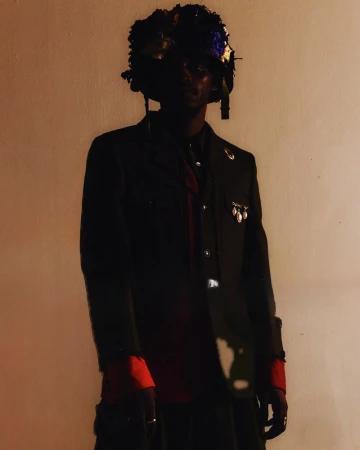Individual in dark jacket with floral headpiece poses in dimly lit setting, creating a visually striking and unique fashion statement.