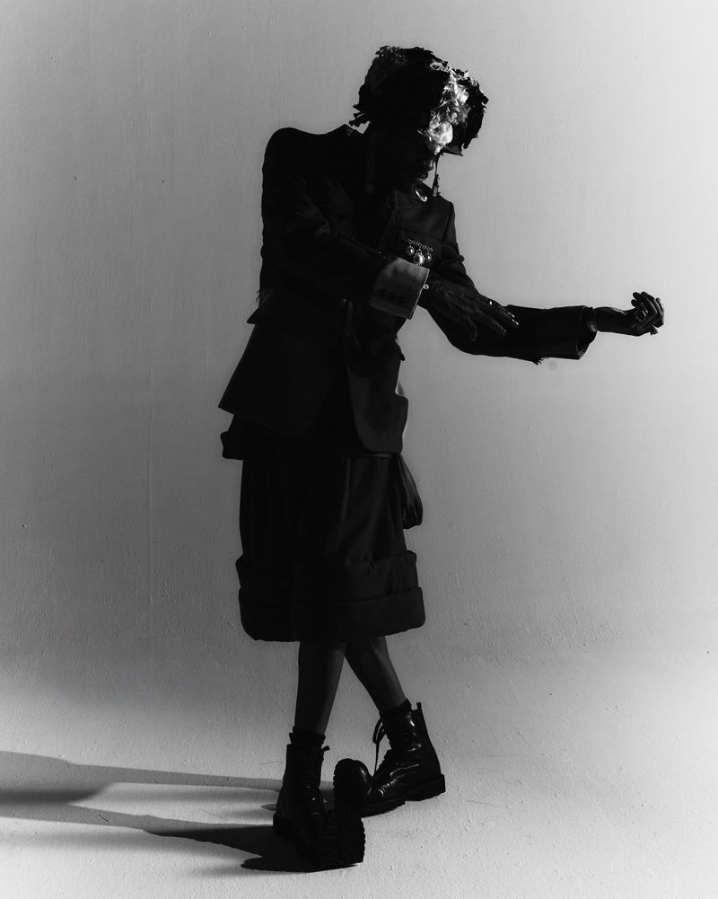 Monochrome studio portrait of a slender adult fashion model in an oversized jacket and boots, creatively styled in dramatic shadows.