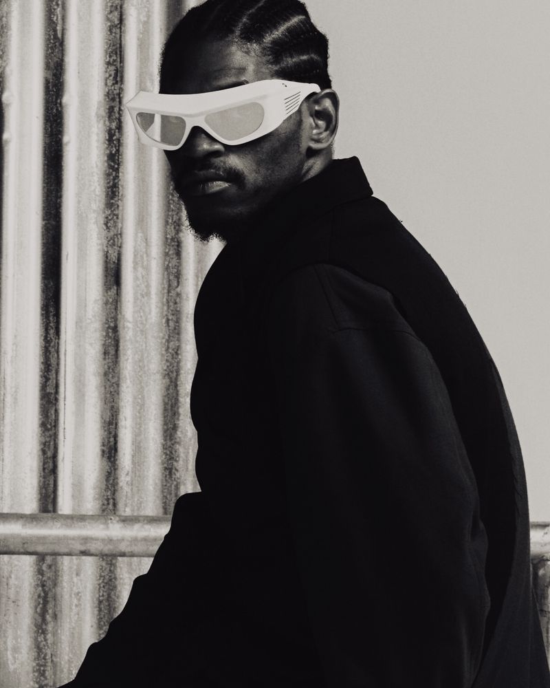 Monochrome portrait of a Black man with stylish sunglasses and a jacket, standing in front of a textured white wall.