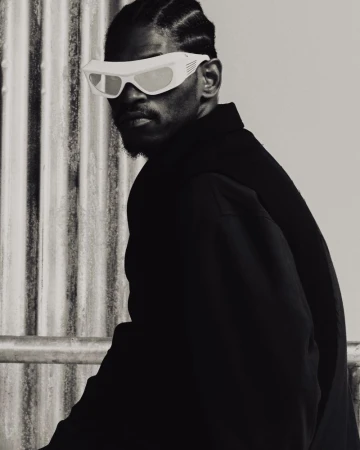 Monochrome portrait of a Black man with stylish sunglasses and a jacket, standing in front of a textured white wall.