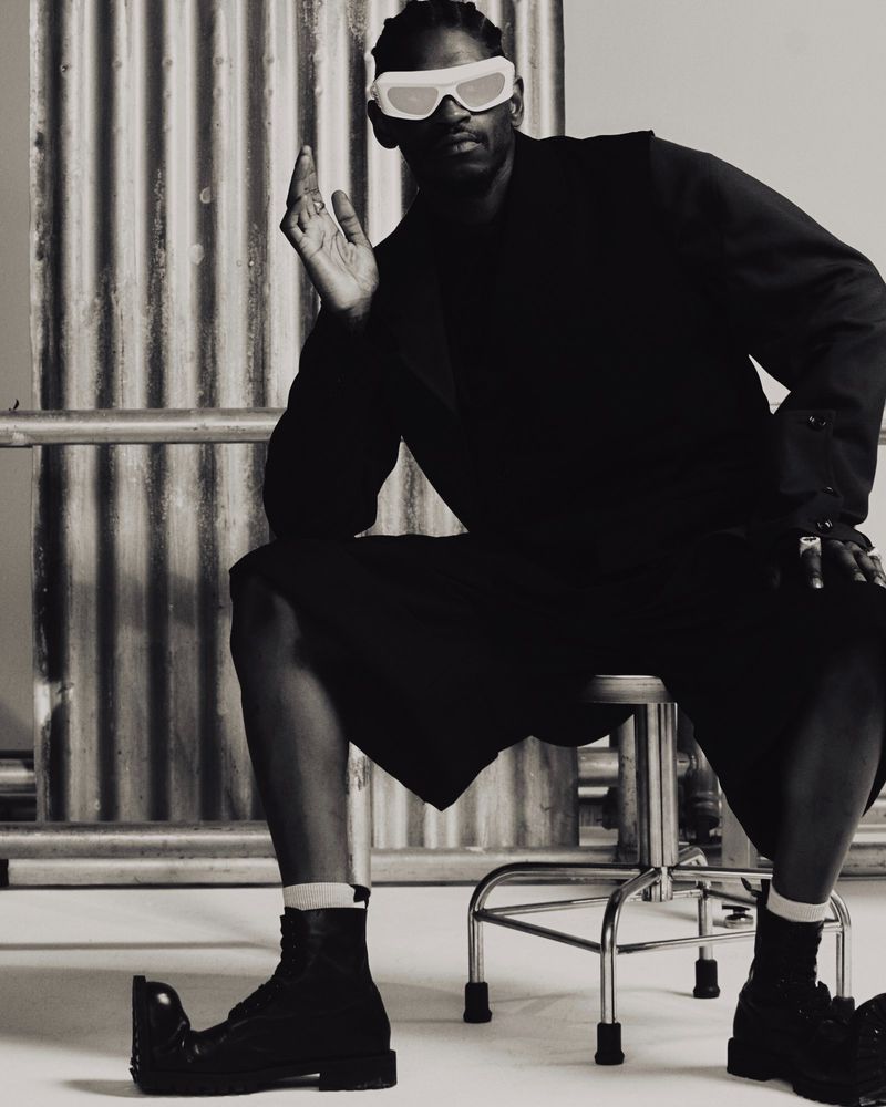 Monochrome studio shot of a Black male model sitting on a stool wearing a black suit, black boots, and white futuristic sunglasses.