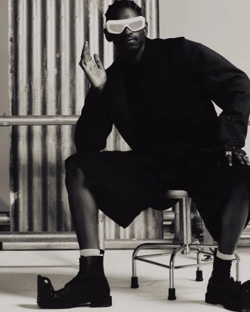 Monochrome studio shot of a Black male model sitting on a stool wearing a black suit, black boots, and white futuristic sunglasses.