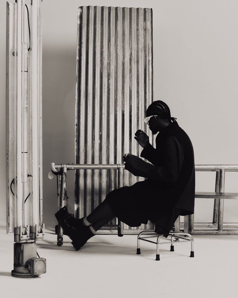 Monochrome studio portrait of a Black male model in dark clothing and futuristic glasses, seated on a chrome stool near metal structures.
