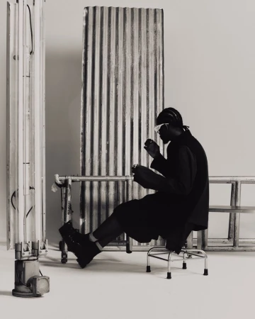 Monochrome studio portrait of a Black male model in dark clothing and futuristic glasses, seated on a chrome stool near metal structures.