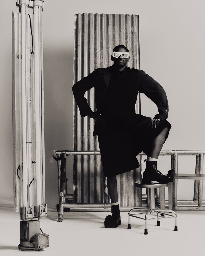 Black and white portrait of an African American adult male in a dark skirt suit and boots, posing against a neutral backdrop with metal props.