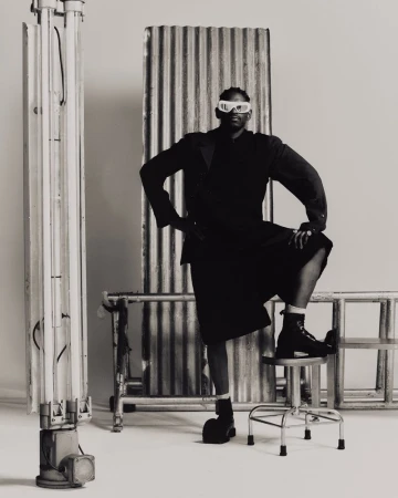 Black and white portrait of an African American adult male in a dark skirt suit and boots, posing against a neutral backdrop with metal props.