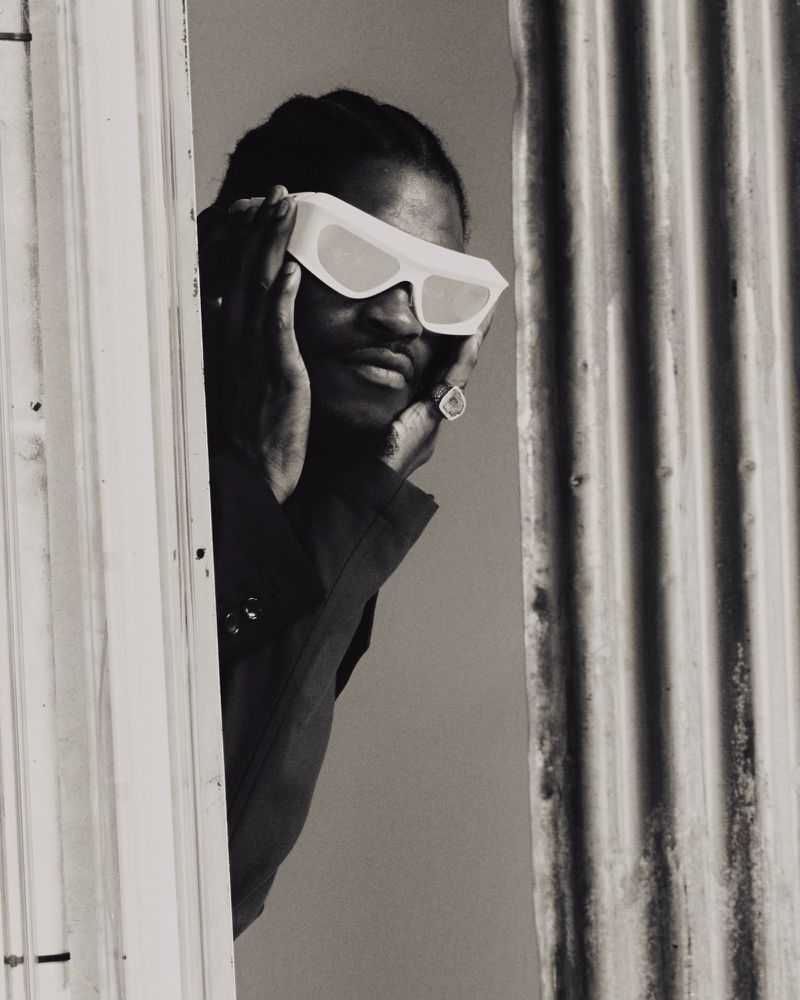Black and white portrait of a male in sunglasses, wearing a dark top, framed by a doorway and corrugated metal background.