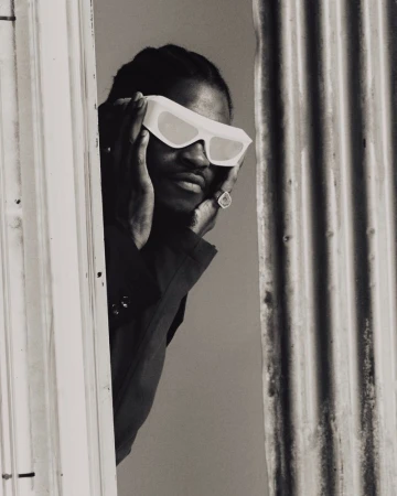 Black and white portrait of a male in sunglasses, wearing a dark top, framed by a doorway and corrugated metal background.