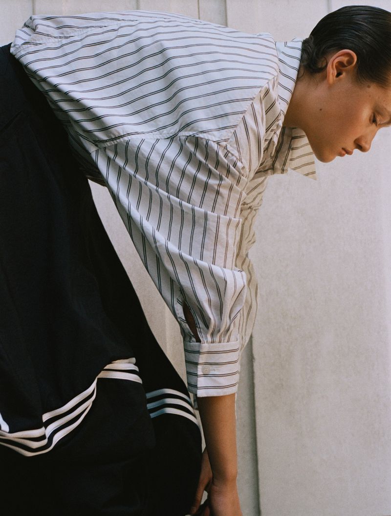 An adult female bends forward wearing a striped shirt and black skirt with white stripes, posing indoors with a neutral facial expression.