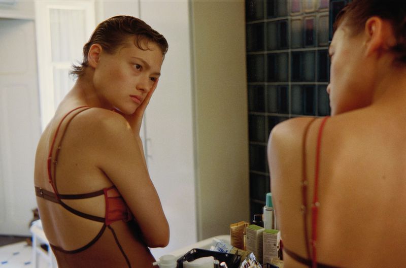 Contemplative woman with wet-look hair gazes into a mirror in a bathroom setting, wearing a stylish brown and red bathing suit.