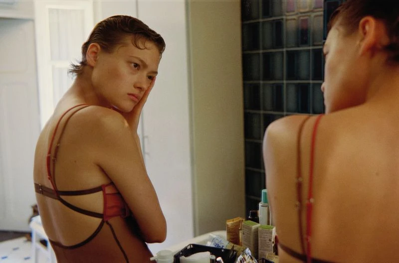 Contemplative woman with wet-look hair gazes into a mirror in a bathroom setting, wearing a stylish brown and red bathing suit.