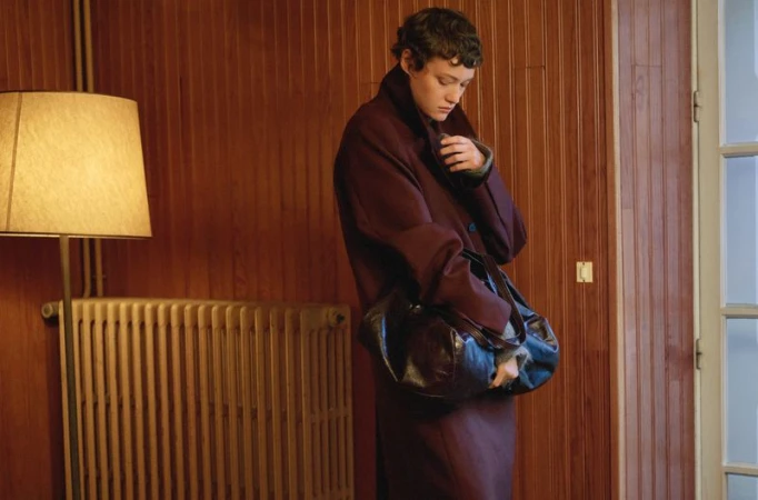 Model Daria Zolotova wearing an oversized burgundy coat and holding a dark leather bag in front of a wood-paneled wall.
