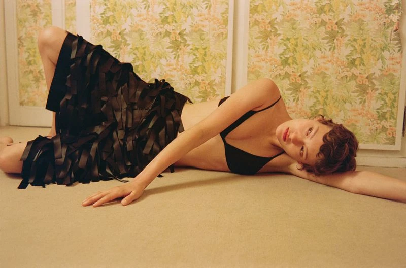 Attractive young woman in a black bra and stylish skirt lies on a beige floor with a floral wallpaper in the background.