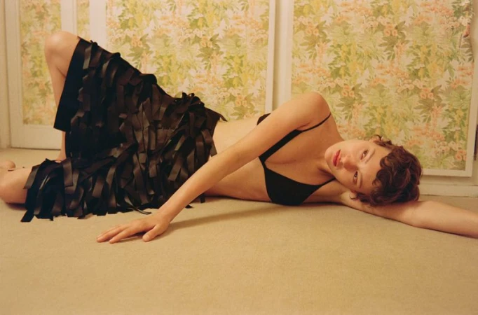 Attractive young woman in a black bra and stylish skirt lies on a beige floor with a floral wallpaper in the background.