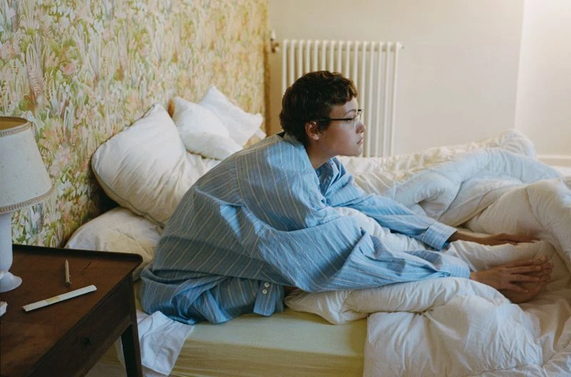 Person in pajamas sitting on a bed with floral wallpaper and a wooden nightstand in an indoor bedroom setting, bathed in soft light.