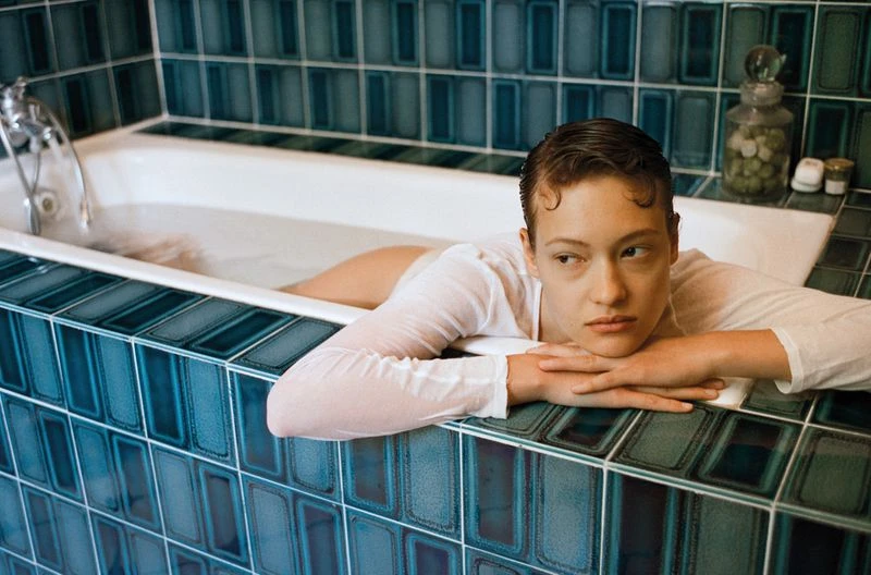 Adult female relaxes in a bathtub with blue tile, wearing a white shirt, looking off to the side with a neutral facial expression.