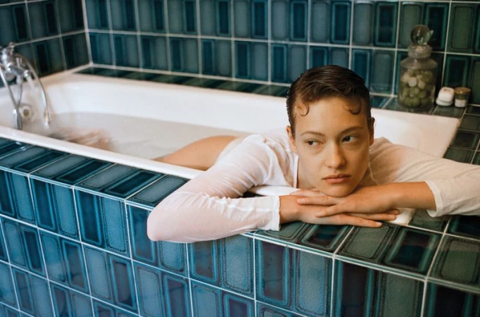 Adult female relaxes in a bathtub with blue tile, wearing a white shirt, looking off to the side with a neutral facial expression.