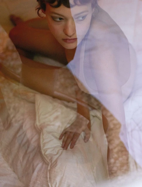 Woman lying on a bed with a white comforter, shot in soft, diffused light creating a dreamy, double exposure effect.